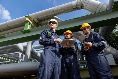 İnşaat alanında tablet ve telsiz kullanarak güvenlik kaskı takan endüstri mühendisleri ekibi takım çalışması, mühendislik planlaması, rafineri denetimi ve saha denetimini temsil ediyor..