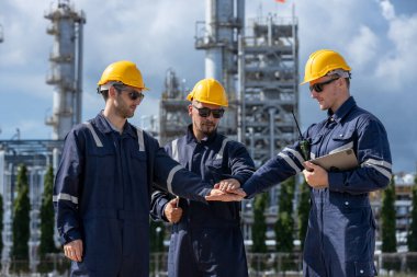 Üç endüstri mühendisi başarıyı neşeli ifadeler ve rafineri tesisinde poz vererek kutluyor, mühendislik endüstrisinde takım çalışması, motivasyon ve başarıyı temsil ediyor..