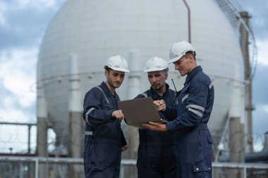 Güvenlik üniforması ve kask giyen bir grup mühendis endüstriyel benzin deposunun önünde dizüstü bilgisayarla projeyi tartışıyor. Takım çalışması kavramı, rafineri denetimi, mühendislik işbirliği güvenliği.