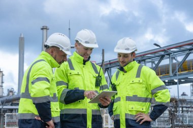 Güvenlik üniforması ve kask giyen bir grup mühendis endüstriyel benzin deposunun önünde projeyi tartışıyor. Takım çalışması kavramı, rafineri denetimi, mühendislik işbirliği güvenliği.