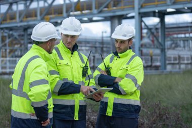 Güvenlik üniforması ve kask giyen bir grup mühendis endüstriyel benzin deposunun önünde projeyi tartışıyor. Takım çalışması kavramı, rafineri denetimi, mühendislik işbirliği güvenliği.