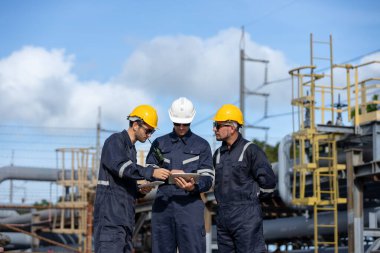 Güvenlik üniforması içindeki bir grup petrol mühendisi boru hattı şantiyesinde dijital tablet kontrolü kullanarak proje planlarını tartışıyor, takım çalışması endüstriyel petrol rafinerisini sembolize ediyor.