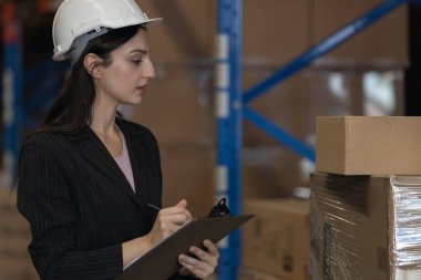 Kendine güvenen kadın depo müdürü elinde bir pano, paletleri inceliyor, lojistik, envanter kontrolü, tedarik zinciri ve endüstriyel depo yönetimini temsil ediyor..