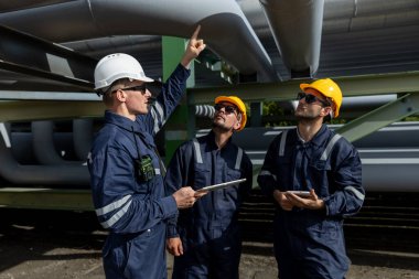 Endüstriyel bir tesiste dijital tablet kullanarak güvenlik donanımı giyen mühendis, büyük boru hatları altında veri ve işlemleri tartışıyor, modern mühendislikteki teknoloji entegrasyonunu sembolize ediyor.