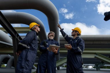 Endüstriyel bir tesiste dijital tablet kullanarak güvenlik donanımı giyen mühendis, büyük boru hatları altında veri ve işlemleri tartışıyor, modern mühendislikteki teknoloji entegrasyonunu sembolize ediyor.