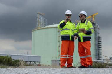 İki erkek mühendis, endüstriyel güvenlik ve teftiş için bulutlu gökyüzünün altında telsiz ve tablet kullanarak, benzin tesisi alanında koordinasyon sağlayan yüksek görüşlü güvenlik teçhizatı giyiyorlar..