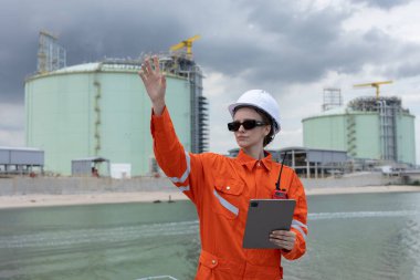 Turuncu üniformalı kadın endüstriyel mühendis, elinde tablet ve telsiz telsiziyle bir benzin santralindeki operasyonları yönetiyor, enerji sektöründeki güvenlik ve iletişim üzerine odaklanıyor..