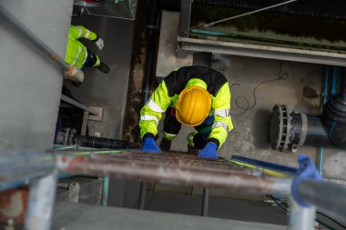 Fabrikanın içinde güvenlik teçhizatı merdiveni tırmanışı yapan sanayi işçisi. Mesleki güvenlik, bakım ve mühendislik kavramı. Yüksek görüş açısına sahip teknisyenlerin üst görünümü.