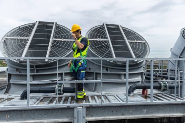 Sanayi teknisyeni, bir imalat tesisinde çatı havalandırma sistemlerini denetliyor, HVAC bakım, mühendislik güvenliği ve endüstriyel altyapı operasyonlarını vurguluyor..