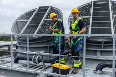 Güvenlik üniformalı iki endüstriyel teknisyen çatıdaki havalandırma ünitelerinde havalandırma sistemi teftişi yapıyor, mühendislik bakımı, güvenlik protokolleri, mekanik operasyonlar üzerinde çalışıyorlar..