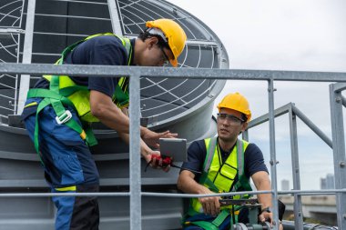 Güvenlik üniformalı iki endüstriyel teknisyen çatıdaki havalandırma ünitelerinde havalandırma sistemi teftişi yapıyor, mühendislik bakımı, güvenlik protokolleri, mekanik operasyonlar üzerinde çalışıyorlar..