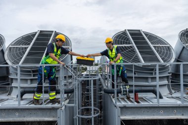 Güvenlik üniforması giyen iki endüstriyel teknisyen çatıdaki AVAC birimlerinden geçen alet kutusundan geçerek takım çalışmasını, mühendislik işbirliğini ve endüstriyel altyapının bakımını sembolize ediyor..