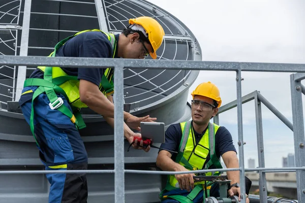 Güvenlik üniformalı iki endüstriyel teknisyen çatıdaki havalandırma ünitelerinde havalandırma sistemi teftişi yapıyor, mühendislik bakımı, güvenlik protokolleri, mekanik operasyonlar üzerinde çalışıyorlar..
