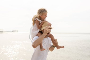 Mutlu bir baba, kumsalda kızını sırtında taşıyor. Hem gülümsüyor hem de yaz tatilinin tadını çıkarıyor. Babalığı, aile bağlarını, ebeveynlik neşesini ve tropikal ortamdaki eğlenceli yaşam tarzını temsil ediyor..