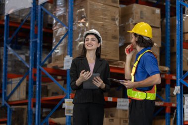 Kadın denetçi ve depo çalışanı lojistik operasyonları tartışmak için tablet ve telsiz kullanıyor. Modern depo ortamında akıllı envanter yönetimiyle takım koordinasyonu.