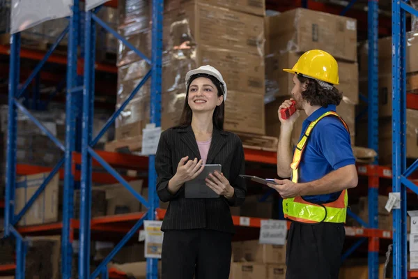 Kadın denetçi ve depo çalışanı lojistik operasyonları tartışmak için tablet ve telsiz kullanıyor. Modern depo ortamında akıllı envanter yönetimiyle takım koordinasyonu.