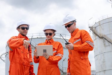 Turuncu güvenlik üniforması ve kasklı mühendisler bir petrol rafinerisinde birlikte çalışıyorlar, takım çalışması, iletişim ve enerji endüstrisinde liderlik ile operasyonlar planlıyorlar..