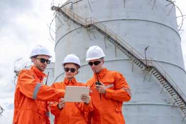 Turuncu güvenlik üniforması ve kasklı mühendisler bir petrol rafinerisinde birlikte çalışıyorlar, takım çalışması, iletişim ve enerji endüstrisinde liderlik ile operasyonlar planlıyorlar..