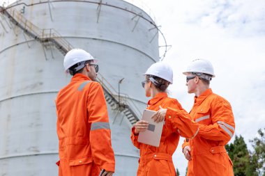 Turuncu güvenlik üniforması ve kask giyen mühendisler petrol depolama tanklarını denetliyor, takım çalışması, saha analizi ve endüstriyel enerji sektöründeki güvenlik prosedürlerine odaklanıyor.