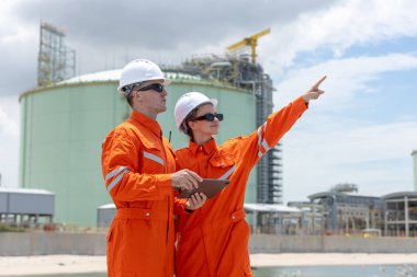 Enerji sektöründe takım çalışması, planlama ve altyapı gelişimini temsil eden, suya yakın bir sanayi bölgesinde proje yönünü inceleyen turuncu güvenlik üniformaları ve miğferleri bulunan mühendisler.