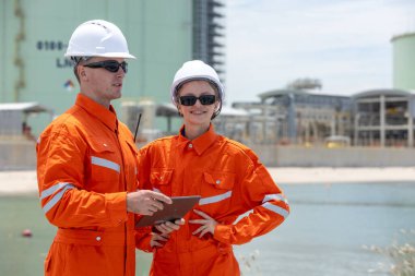 Su yakınlarındaki bir sanayi bölgesinde proje planlarını tartışan turuncu güvenlik üniformaları ve miğferleri mühendisi, takım çalışması, iletişim, enerji altyapısı geliştirme çalışmalarını vurguluyor.