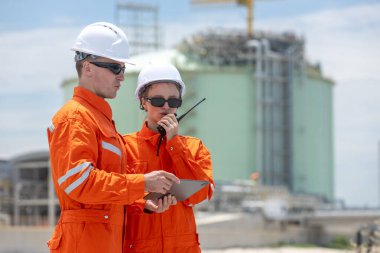 Turuncu güvenlik üniformalı mühendisler, modern endüstriyel santralde bir tablet ve telsiz kullanıyorlar. Enerji sektöründe iletişim, koordinasyon, alan planlaması gösteriyorlar..