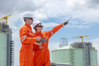 Enerji sektöründe takım çalışması, planlama ve altyapı gelişimini temsil eden, suya yakın bir sanayi bölgesinde proje yönünü inceleyen turuncu güvenlik üniformaları ve miğferleri bulunan mühendisler.