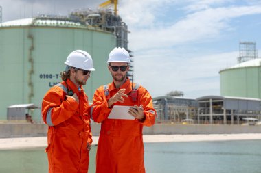 Turuncu güvenlik üniforması ve miğferli iki mühendis limandaki bir sanayi bölgesinde verileri tartışıyor, takım çalışması, analiz ve modern enerji operasyonlarını temsil ediyorlar..