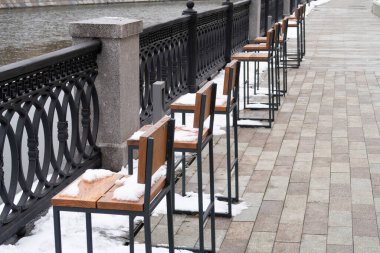 Kanal kenarındaki metal çitlerin yanında karla kaplı ahşap koltuklu bir sıra sandalye.