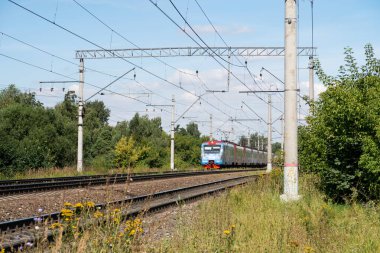 Kablolar ve beton direklerle raylar, yaz yeşillikleri arasında mavi tren.