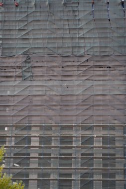 İnşaat halindeki yüksek bir binanın ön cephesi çatı örgüsü ve arka planla kaplıdır.