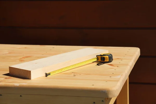 On the light surface of a wooden table there is a scrap of a board and a tape measure for carpentry work