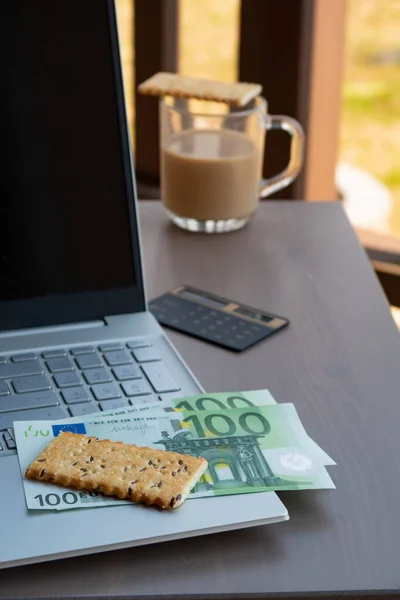 There is coffee on the table in a glass glass with cookies, a laptop and a calculator with European banknotes against the background of a summer cottage, working remotely