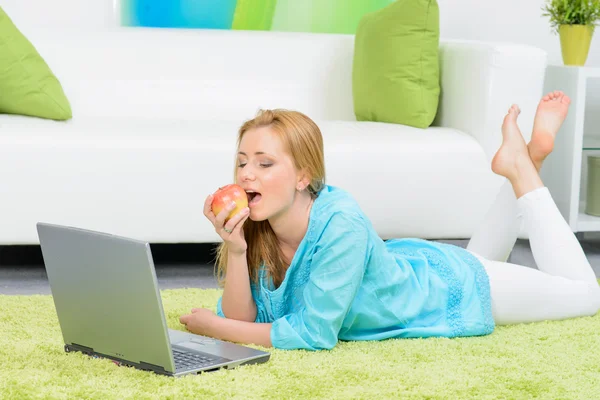 Красивая женщина дома лежит на полу с ноутбуком едят яблоко — стоковое фото