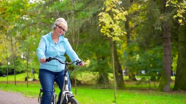 快乐的老年妇女戴着耳机 骑着自行车 听着音乐有声读物 骑复古自行车的老骑自行车的妇女追求积极的自然生活方式 漫步秋天的森林或城外的城市公园 — 图库视频影像