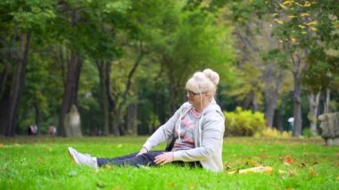 Emekli yaşlı bir kadının portresi çimenlikte paspas üzerinde egzersiz yapıyor yaşlı bir kadın doğada yoga pozisyonu alıyor. Emeklilikte aktif sağlıklı yaşam tarzı. Gözlüklü ve spor kıyafetli büyükanne.