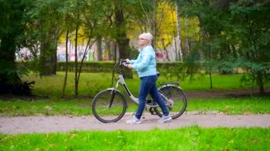 Gözlüklü, sıradan kıyafetleri olan mutlu bir son sınıf kadını bisikletle parkta yürüyor. Motosikletli yaşlı kadın bisikletçi doğada aktif yaşam tarzını takip ediyor. Sonbahar ormanlarında ya da şehir parkında bir yürüyüş.