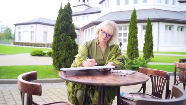 昼間は夏のテラスでシニア女性アーティストがペイント 年配の女性デザイナーがテーブルで路上生活をしている 魅力的な老女が描画し 彼女の好きな趣味や仕事を屋外 — ストック動画