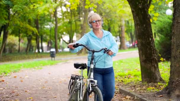 Mulher Sênior Feliz Fones Ouvido Caminha Parque Com Uma Bicicleta — Vídeo de Stock