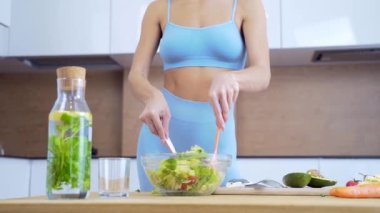 Mutfakta sağlıklı yemek pişiren spor giyinen genç bir fitness kadını. Sebze salatası hazırlayın. Diyet kahvaltı hazırlayan çekici bir kız. Atletizm, yemekleri cam tabakta veya kasede karıştırır.