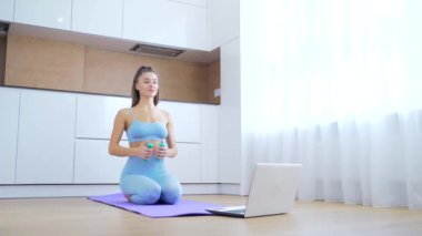 Spor kıyafetli genç çekici bir kadın mutfakta yerde oturan dizüstü bilgisayarının önünde halterlerle online egzersiz yapıyor. Kız evde karantina eğitimi alarak spora uzak mesafeden gidiyor.