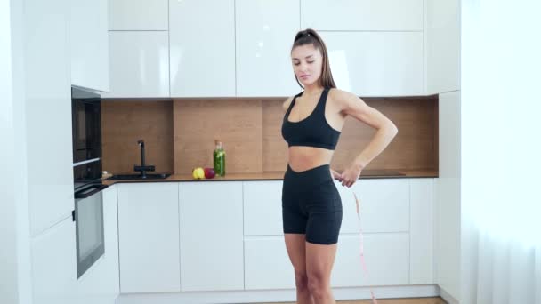 在厨房里 穿着运动服的身材纤细 腰身纤细的年轻健身女子用带子测量腰围 有魅力的女运动员在家里做一些测量 — 图库视频影像