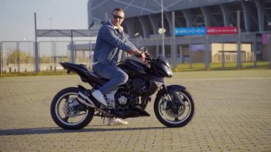 Uban arka planında kendine güvenen şık genç erkek motorcunun portresi. Güneş gözlüklü genç bir adam hız yapan bir motosiklete ya da bisiklete biner ve kameraya bakar. Dışarıda kot elbiseli bir adam var.