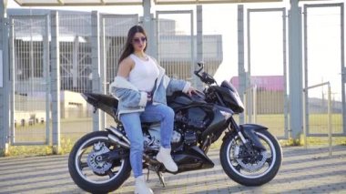 Spor motosikletinde oturan çekici, genç ve şık bir kızın portresi. Kot elbiseli, güneş gözlüklü, şehir arka planında bisikleti olan seksi bir moda kadını. Kameraya bakıp poz vermek.