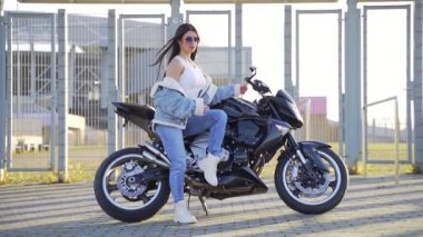 Spor motosikletinde oturan çekici, genç ve şık bir kızın portresi. Kot elbiseli, güneş gözlüklü, şehir arka planında bisikleti olan seksi bir moda kadını. Kameraya bakıp poz vermek.