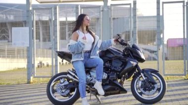 Spor motosikletinde oturan çekici, genç ve şık bir kızın portresi. Kot elbiseli, güneş gözlüklü, şehir arka planında bisikleti olan seksi bir moda kadını. Kameraya bakıp poz vermek.
