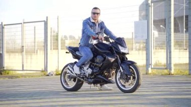 Uban arka planında kendine güvenen şık genç erkek motorcunun portresi. Güneş gözlüklü genç bir adam hız yapan bir motosiklete ya da bisiklete biner ve kameraya bakar. Dışarıda kot elbiseli bir adam var.