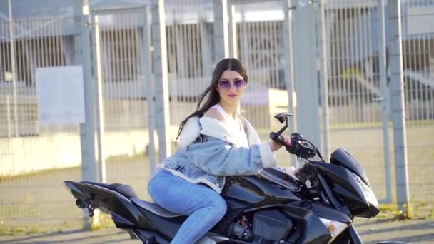スポーツバイクに座って魅力的な若い大人のスタイリッシュな女の子の肖像画 都会的な街の背景に自転車でデニム服やサングラスでセクシーなファッション女性 カメラを見てポーズをとって — ストック動画
