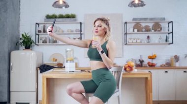 Formda genç bir kadın mutfak içinde bir bardak taze sebze suyuyla fotoğraf için poz veriyor. Kameralı telefonun önünde sağlıklı içkiler içen mutlu kadın. Akıllı telefondan video çeken güzel kız.