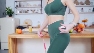 Kapatın. Genç fitness kadını ince ince bir atletik figür mutfakta bir mezura ile bel çevresini ölçer. Çekici kız evin arka planındaki meyvelerin ölçüsünü alıyor.